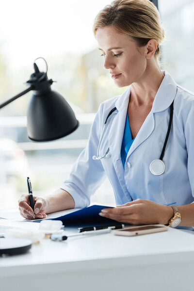 confident adult female doctor in white coat with stethoscope writing in clipboard at table in office 