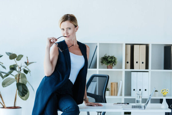 beautiful adult businesswoman holding eyeglasses and posing on table with laptop in office 