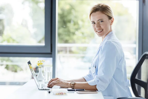 Lächelnde Ärztin Weißen Mantel Blickt Die Kamera Und Arbeitet Laptop — kostenloses Stockfoto