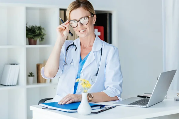 Médico Femenino Adulto Sonriente Ajustando Anteojos Mesa Con Computadora Portátil —  Fotos de Stock