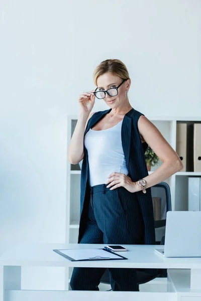 Erwachsene Geschäftsfrau Justiert Brille Und Blickt Kamera Tisch Mit Laptop — kostenloses Stockfoto