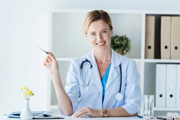 Gülümseyen Kadın Doktor Ofisinde Masada Termometre Gösterilen Beyaz Paltolu — Stok fotoğraf