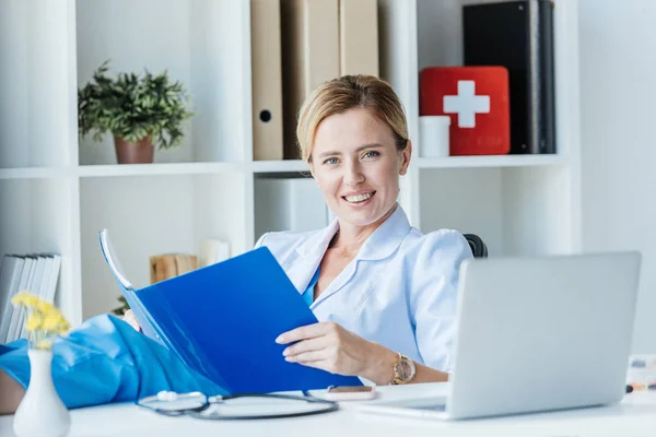 Mujer Feliz Médico Capa Blanca Con Portapapeles Mirando Cámara Mesa — Foto de stock gratis