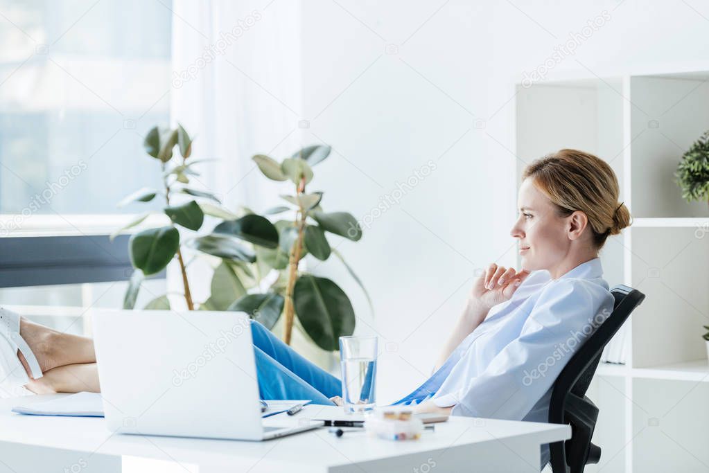side view of thoughtful female doctor with legs on table sitting in office 