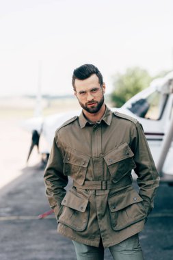 stylish man in green jacket and eyeglasses posing with hands in pockets near airplane  clipart