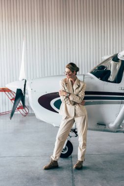fashionable woman in sunglasses and jacket posing near aircraft in hangar clipart