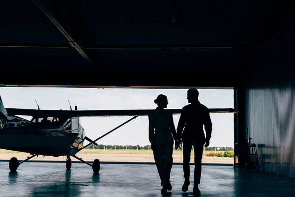 rear view of silhouettes of young couple walking near airplane in hangar 