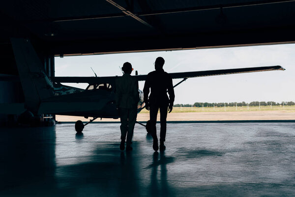 rear view of silhouettes of stylish couple walking near airplane in hangar 