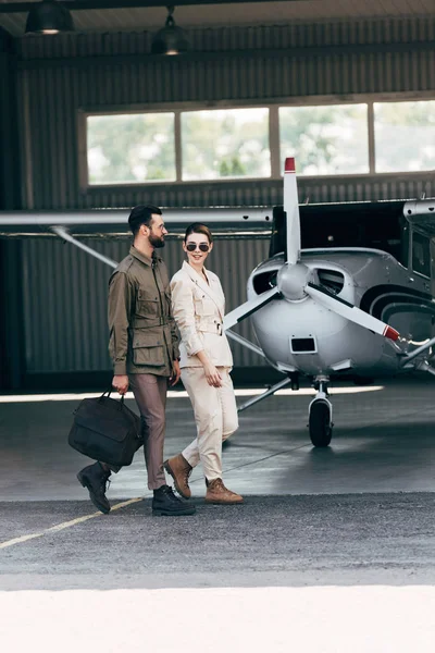 stylish man carrying bag and walking with girlfriend near hangar with plane