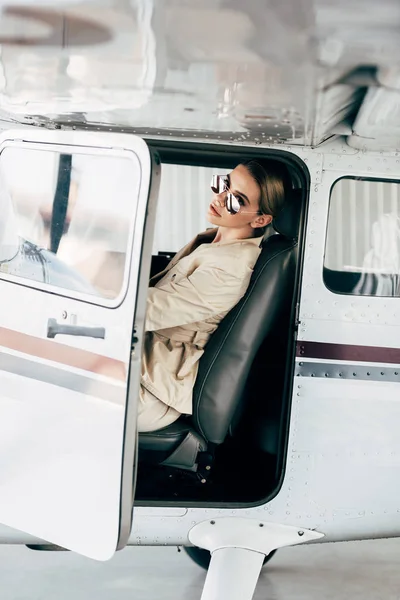 时尚的年轻女子在太阳镜和夹克坐着 看着相机在飞机上 — 免费的图库照片