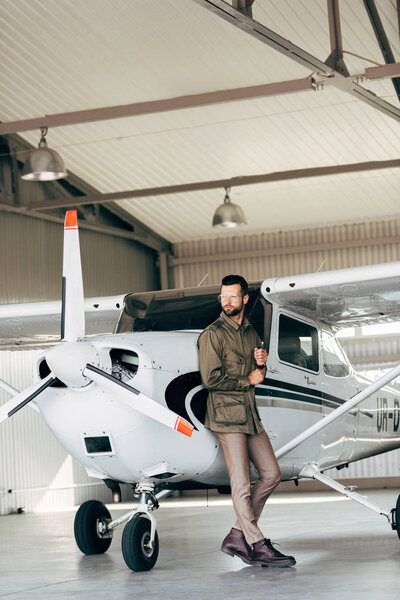 fashionable young man in green jacket and eyeglasses posing near airplane 