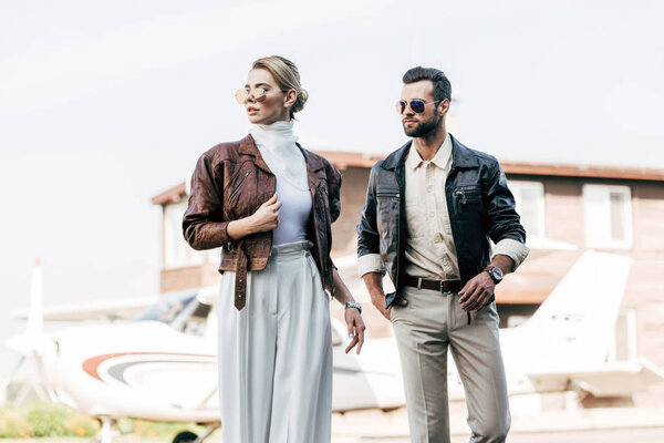 stylish couple in sunglasses and leather jackets standing near airplane