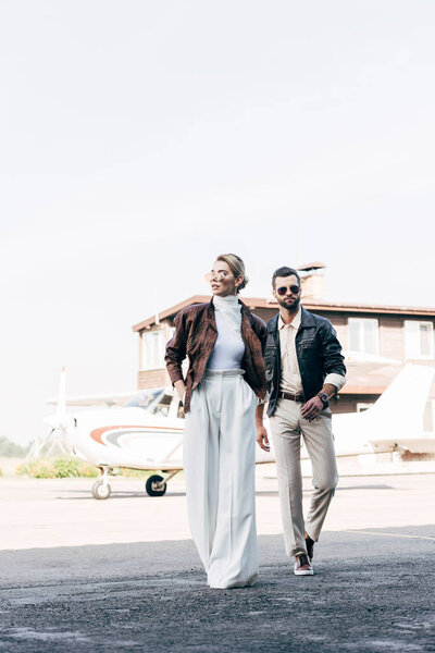 young stylish couple in sunglasses and leather jackets walking near airplane