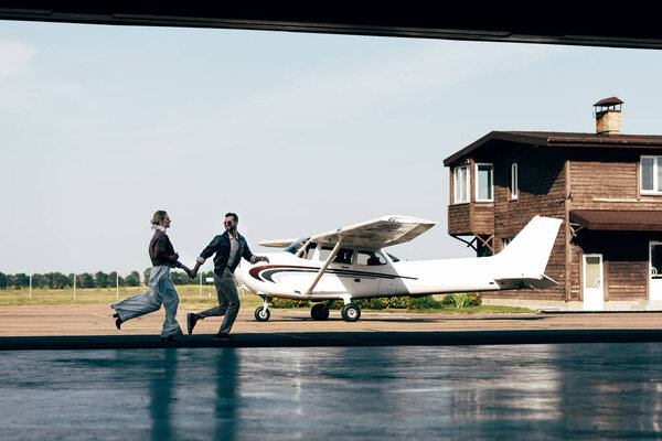 distant view of stylish young couple in leather jackets holding hands and running near plane 