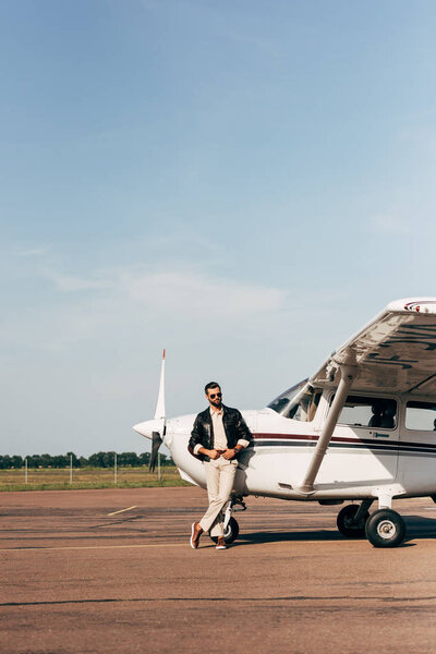 fashionable male pilot in leather jacket and sunglasses posing near plane