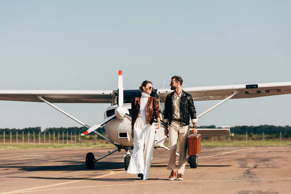 stylish young couple in leather jackets and sunglasses walking with retro suitcase near airplane 