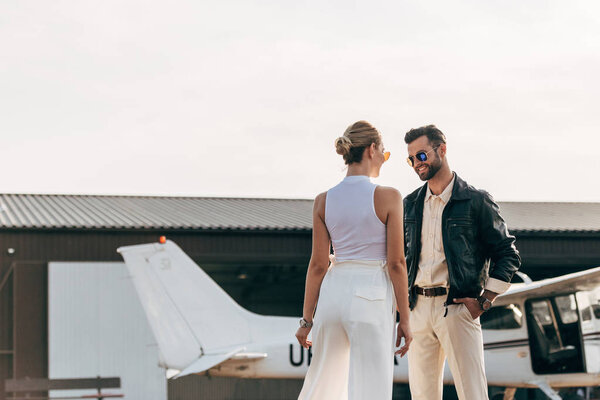 rear view of stylish woman talking to smiling boyfriend in leather jacket and sunglasses near plane 