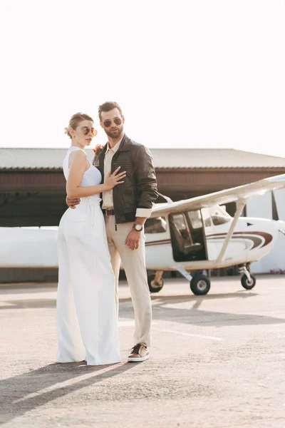 handsome young man in leather jacket and sunglasses embracing attractive girlfriend near plane