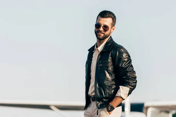 年轻英俊的男性飞行员在皮革夹克和太阳镜摆在飞机附近 — 图库照片