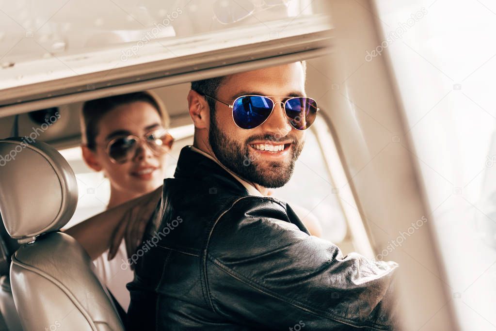 happy male pilot in sunglasses and leather jacket looking at camera while his girlfriend sitting near in cabin of airplane 