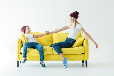 side view of mother and son in burgundy hats holding hands on yellow sofa on white clipart