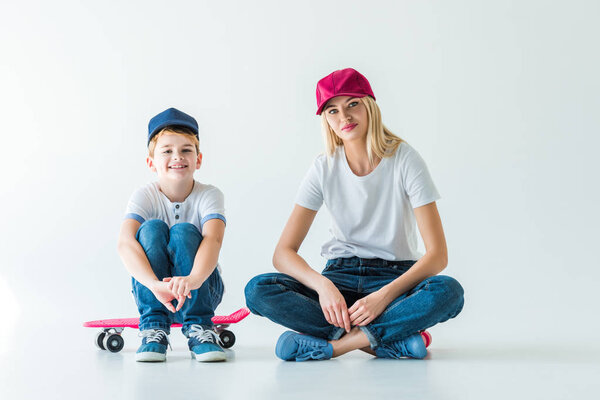 cheerful mother and son in caps sitting on skates and looking at camera on white
