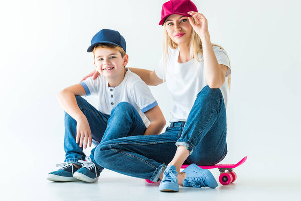 mother and son in caps sitting on skates, hugging and looking at camera on white