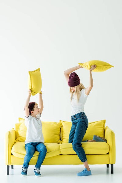 mother and son in casual clothes having fun and fighting with pillows on yellow sofa on white