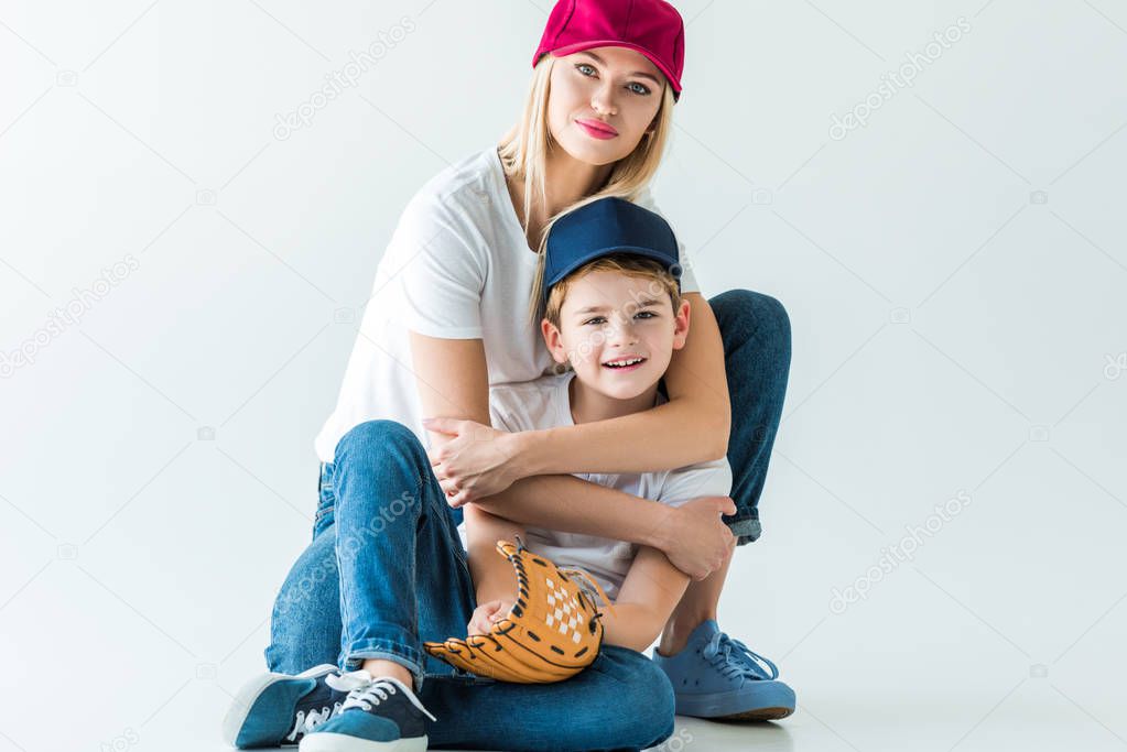 smiling mother hugging son and sitting on floor with baseball glove on white