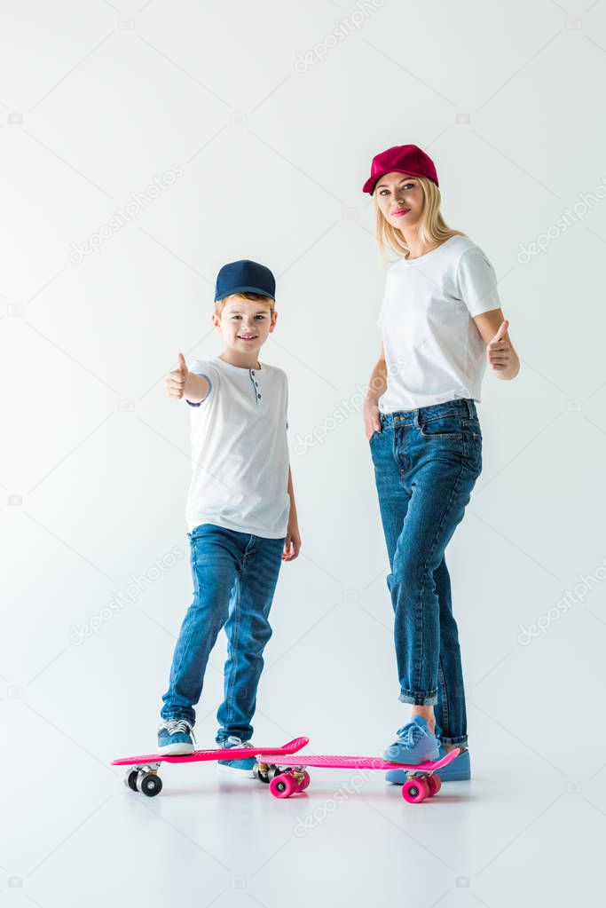 smiling mother and son in caps standing near skates and showing thumbs up on white