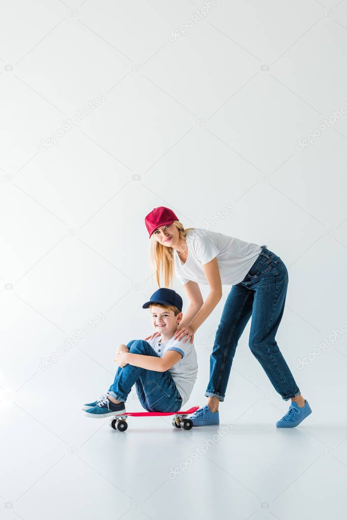 happy mother in red cap pushing son on skate on white