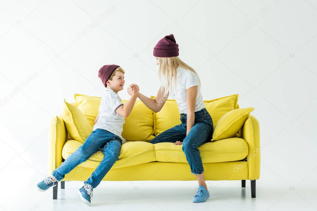 mother and son in burgundy hats arm wrestling on yellow sofa on white