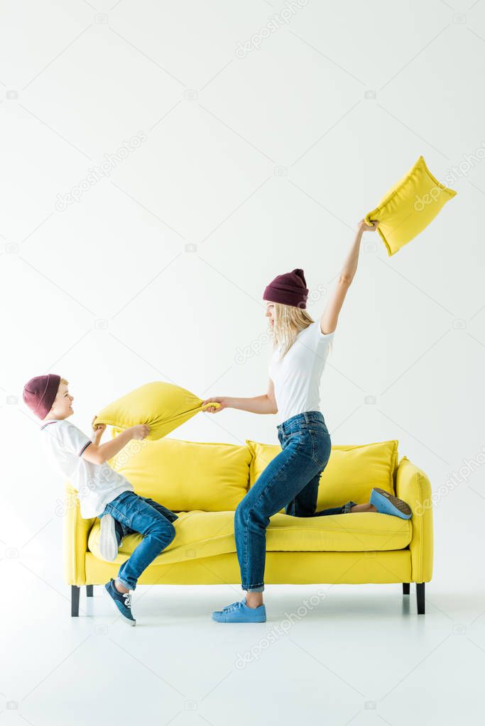 mother and son in burgundy hats having fun and fighting with cushions on yellow sofa on white