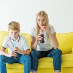Mãe e filho bebendo refrigerante saboroso com palhas no sofá amarelo isolado no branco