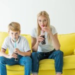 Madre e hijo bebiendo sabroso refresco con pajitas en sofá amarillo aislado en blanco