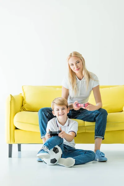 微笑的母亲和儿子玩视频游戏黄色沙发上白色 足球在地板上 — 图库照片
