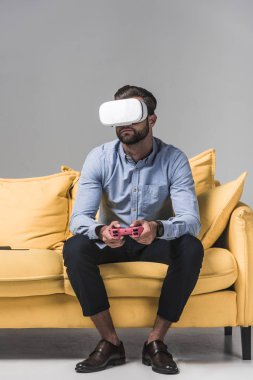 adam gri sarı kanepe üzerinde joystick ve vr kulaklık ile bilgisayar oyunu oynamak