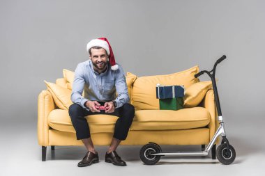 mutlu Noel hediyeleri ve skuter Grey ile kanepede otururken joystick ile bilgisayar oyunu oynamak santa şapka erkekte