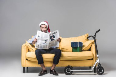 yakışıklı adam santa şapka Noel hediyeleri ve skuter Grey ile sarı kanepede otururken iş gazete okuma