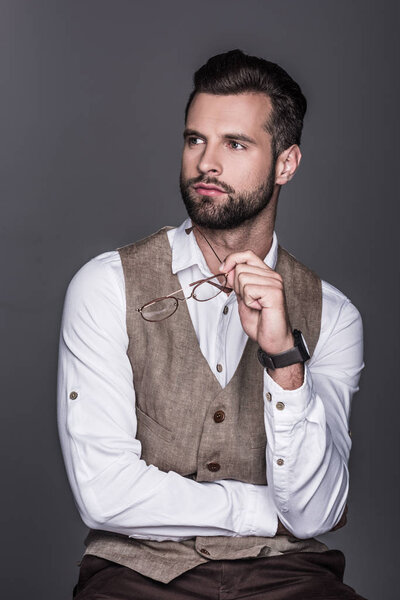 portrait of handsome bearded man holding glasses and posing in waistcoat, isolated on grey 
