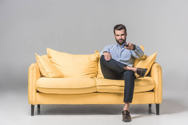 handsome smiling man with remote control watching TV and sitting on yellow sofa on grey