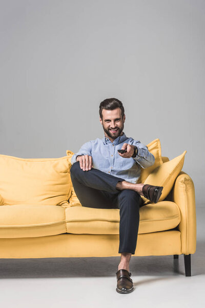 cheerful bearded man with remote control watching TV and sitting on yellow sofa on grey