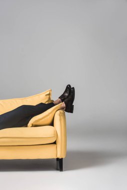 cropped view of male legs lying on yellow sofa on grey clipart