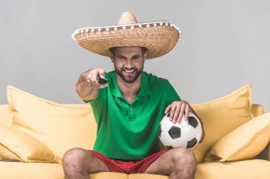 Meksika sombrero futbol topu ve uzaktan kumandayla Grey sarı kanepede otururken maçı izlerken erkekte gülümseyen  
