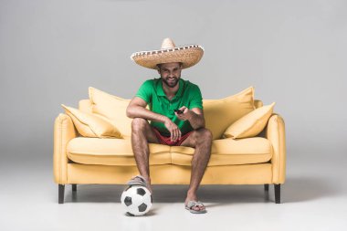 futbol topu ve uzaktan kumandayla Grey sarı kanepede otururken maçı izlerken sombrero Meksika yakışıklı adam  