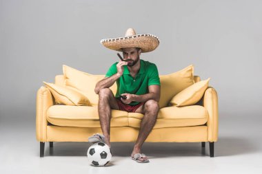 ciddi adam Meksika sombrero izlerken futbol maç puro, top ve uzaktan kumanda üzerindeki gri sarı kanepede otururken  