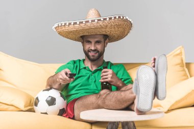 smiling man in mexican sombrero watching football match while sitting on yellow sofa with ball, bottle of beer and remote control on grey   clipart