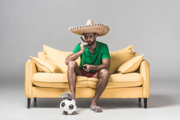 serious man in mexican sombrero watching soccer match while sitting on yellow sofa with cigar, ball and remote control on grey  