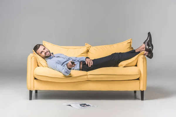 Smiling man with remote control watching TV and lying on sofa with newspaper on floor, on grey — Stock Image Smiling Man Remote Control Watching Lying Sofa Newspaper Floor Grey Stock Picture