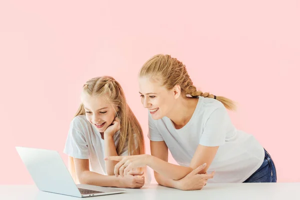 smiling mother and daughter in white t-shirts pointing and looking at laptop together isolated on pink